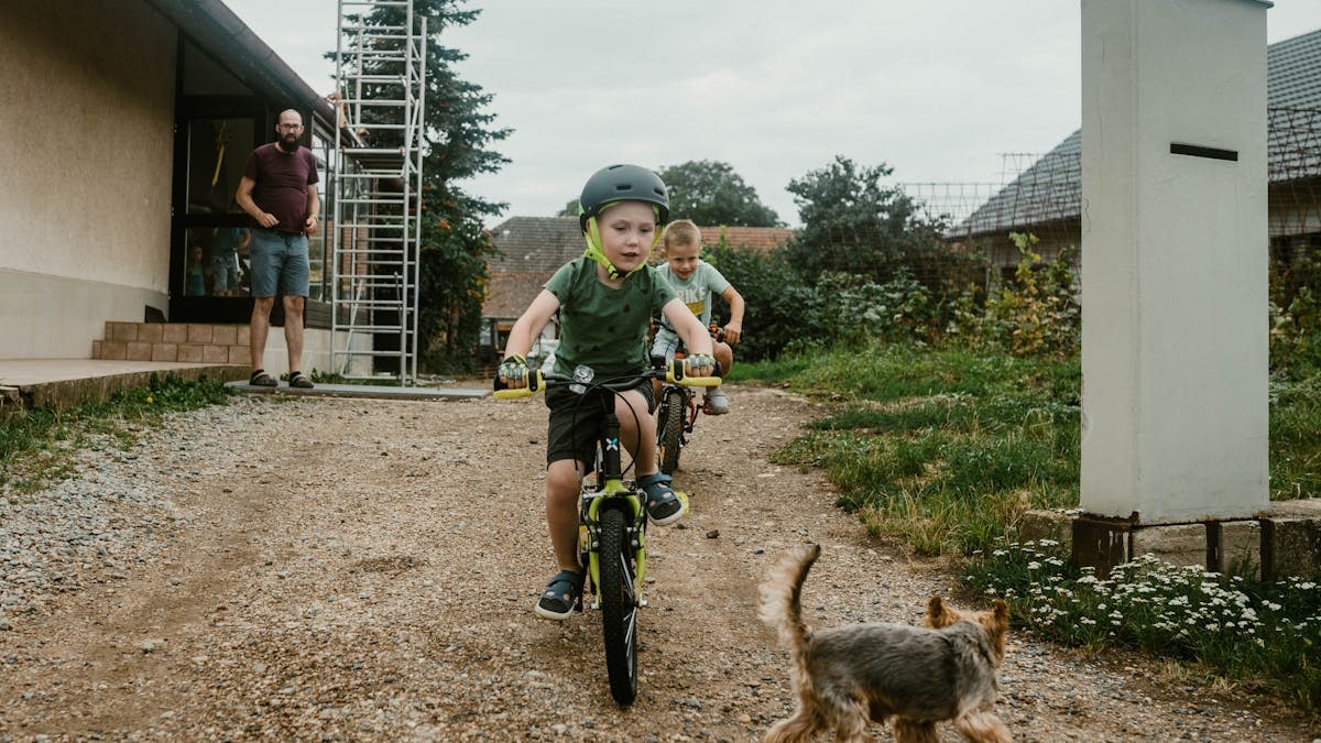 Two kids cycling on a dirt path with a small dog chasing them, adult supervising nearby.